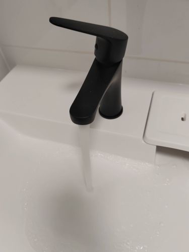 Изображение показывает современный черный смеситель, установленный на белой раковине в ванной комнате. Вода течет из крана, создавая легкую пену на поверхности.