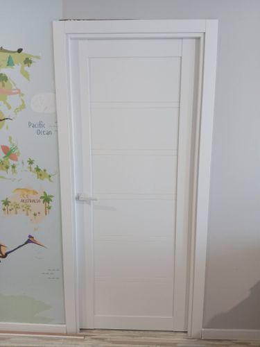 Установлена новая белая дверь с ручкой, закреплена коробка, отрегулированы петли, обеспечена плавность закрывания, добавлены наличники, завершена отделка проема, проверена функциональность.