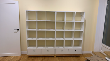 Собран белый стеллаж с множеством квадратных ячеек и тремя нижними ящиками. Конструкция установлена на деревянный пол у стены, рядом с дверью и окном.