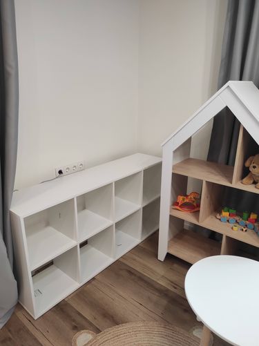 Собран и установлен белый стеллаж с девятью отделениями, аккуратно размещённый в углу комнаты. Все детали надежно закреплены, обеспечивая устойчивость и функциональность конструкции.