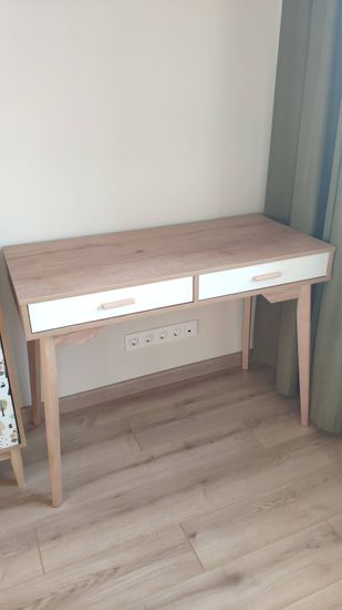 Собран деревянный стол с двумя выдвижными ящиками. Установлены ножки, закреплены ручки, проверена устойчивость. Готовое изделие стоит на светлом полу у стены с розетками.