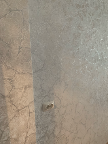 Стены оклеены обоями с текстурным узором, создающим эффект мрамора. Поверхность ровная, стыки аккуратные, розетка интегрирована без зазоров. Работа завершена качественно.