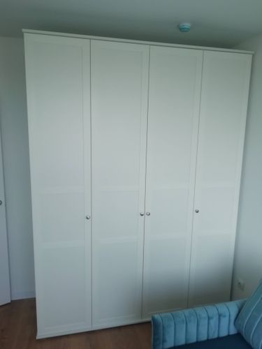 Собран белый шкаф с четырьмя дверцами, установлен в комнате. Все детали надежно закреплены, дверцы ровно висят, ручки прикручены. Шкаф стоит на деревянном полу, рядом голубой диван.