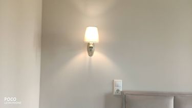 Изображена установленная настенная лампа на светлой стене, рядом с выключателем и розеткой. Светильник излучает мягкое освещение, создавая уютную атмосферу в комнате.