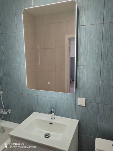 Зеркало аккуратно установлено на стену ванной комнаты, выровнено по уровню, надежно закреплено крепежными элементами, подключена подсветка для удобства использования.