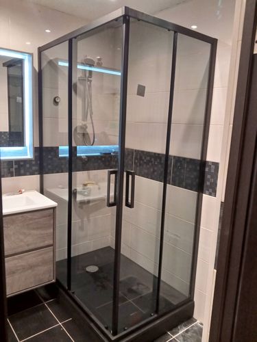 Установлен стеклянный уголок с черными рамами, герметично закреплен на плитке. Подключены водопроводные элементы, смонтированы двери, проверена функциональность.