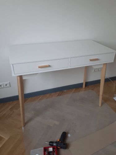 Собран белый стол с двумя ящиками и деревянными ножками. Все детали аккуратно соединены, конструкция устойчива. Инструменты использованы для точной подгонки элементов.