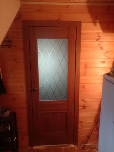 Изображена установленная дверь-гармошка с деревянной рамой и стеклянной вставкой, украшенной узором. Она гармонично вписана в интерьер из деревянных панелей.