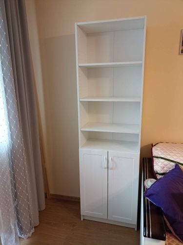 Собрана конструкция с полками и дверцами, установлена в углу комнаты. Стеллаж устойчивый, выполнен из светлого материала, подходит для хранения вещей. Завершённый проект.