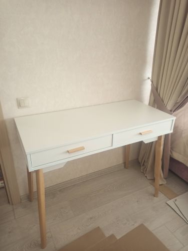 Собран белый стол с двумя выдвижными ящиками и деревянными ножками. Установлены ручки, выровнена поверхность, проверена устойчивость. Готов к использованию в интерьере комнаты.