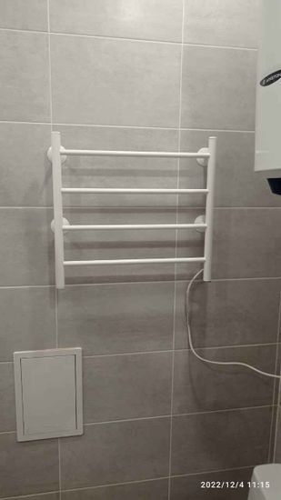 На стене ванной комнаты закреплен электрический полотенцесушитель. Устройство подключено к электросети, кабель аккуратно уложен. Установка завершена, прибор готов к использованию.