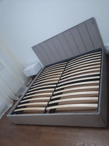 Изображение показывает кровать с поднятым основанием и ламелями, стоящую на деревянном полу рядом с тумбой и занавесками.