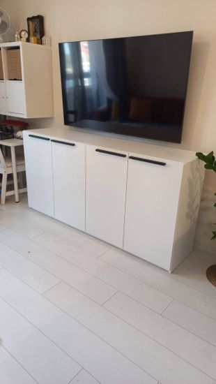 Собрана белая тумба с четырьмя дверцами и черными ручками. Установлена на светлом полу, рядом с телевизором. Все элементы аккуратно подогнаны, конструкция устойчива и надежна.