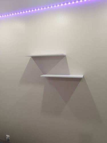 На стене установлены две белые полки, закрепленные ровно и надежно. Верхняя часть стены украшена светодиодной подсветкой, создающей мягкое освещение.