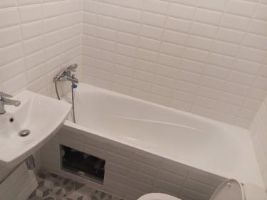 Изображение ванной комнаты с белой плиткой, ванной, раковиной и смесителем. Видно открытое отверстие под ванной, вероятно, для доступа к коммуникациям.