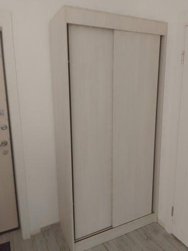 Изображение шкафа с раздвижными дверями в светлом интерьере. Стены белые, пол серый. Шкаф установлен в углу рядом с дверью.