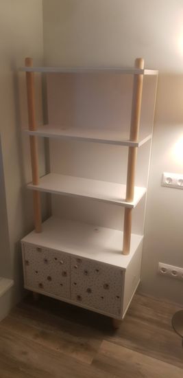 Собран белый стеллаж с деревянными стойками и двумя полками. Установлен на полу, закреплен у стены. Нижняя часть оборудована шкафчиком с узорчатыми дверцами.