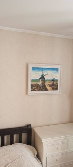 На стене закреплена картина в белой раме с изображением мельниц. Работа выполнена аккуратно, крепление надежное, поверхность осталась без повреждений.