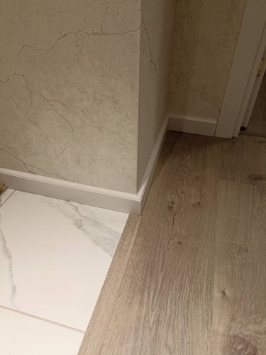 Установлен белый плинтус, аккуратно прилегающий к стене и полу, создавая плавный переход между плиткой и ламинатом. Работа выполнена точно, без зазоров и неровностей.