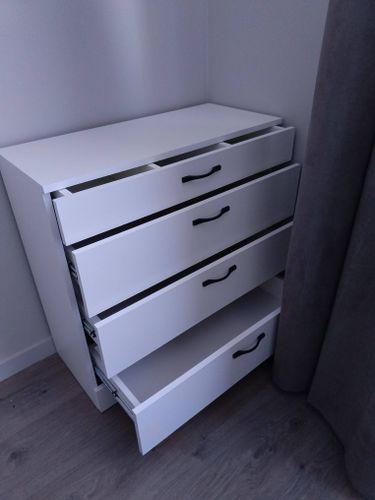 Собран белый комод с четырьмя выдвижными ящиками и черными ручками. Установлены направляющие, закреплены все детали, обеспечена устойчивость конструкции.