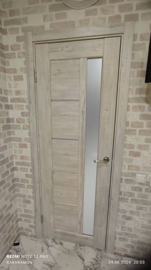 Установлена светлая межкомнатная дверь с вертикальным стеклом, закреплена в проеме, оснащена ручкой и петлями. Завершены работы по монтажу в кирпичном интерьере.