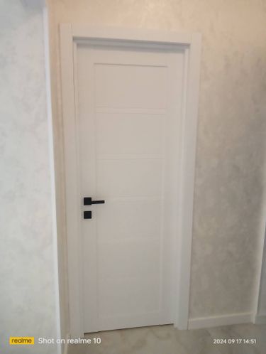 Изображена установленная белая дверь-гармошка с черной ручкой, аккуратно встроенная в дверной проем, гармонично сочетающаяся с интерьером светлой комнаты.