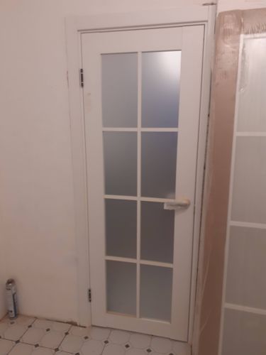 Установлена новая белая дверь с матовыми стеклянными вставками, закреплены петли, ручка, отрегулировано положение в проеме, обеспечена плавная работа, завершены отделочные работы.