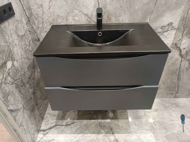 Установлена новая раковина с черным краном на серую тумбу, выполнено подключение к водопроводу и канализации, проверена герметичность всех соединений, завершено тестирование работоспособности.