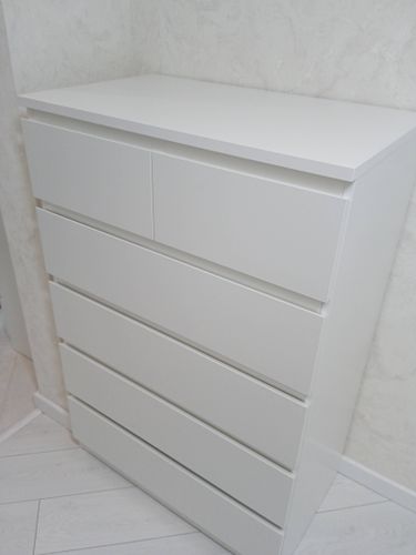 Собран белый комод с шестью ящиками, аккуратно установлен в углу комнаты. Все детали надежно закреплены, фасады ровные, конструкция устойчивая, поверхность гладкая, без дефектов.