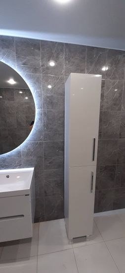 Изображение ванной комнаты с круглым зеркалом с подсветкой, белой раковиной и высоким шкафом на фоне серой плитки.