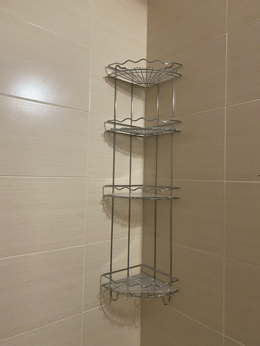 Изображена установленная угловая металлическая полка с тремя ярусами на кафельной стене ванной комнаты. Полка надежно закреплена, обеспечивает удобное хранение.