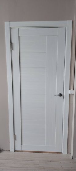 Установлена новая деревянная дверь с петлями и ручкой. Проведены работы по монтажу в проем, закреплены наличники, проверена плавность открывания и закрывания. Завершено оформление.