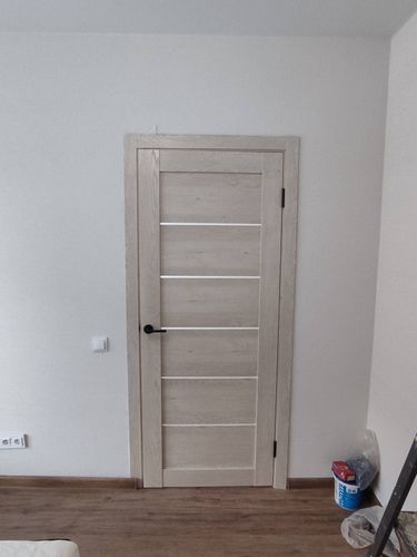 Установлена светлая дверь с ручкой и петлями, закреплена в проеме, отрегулирована по уровню, добавлены наличники, обеспечена плавная работа, завершена отделка.