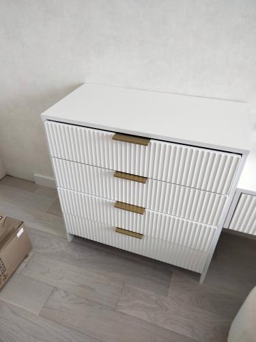 Собран белый комод с четырьмя ящиками и золотыми ручками. Установлены все элементы, обеспечена устойчивость и функциональность. Завершён монтаж на светлом полу.