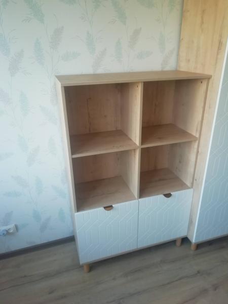 Собран деревянный стеллаж с четырьмя полками и двумя нижними закрытыми секциями. Установлены ножки, дверцы закреплены. Готовая конструкция стоит на полу, прилегает к стене.