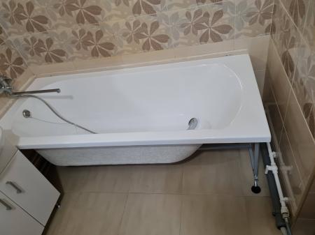 Изображение ванной комнаты с установленной новой белой ванной, подключенной к водопроводу, на фоне кафельной стены с узором. Пол выложен плиткой, трубы аккуратно уложены.