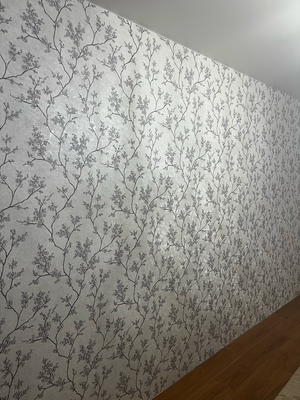 На стену аккуратно наклеены обои с ветвями и цветами, создавая гармоничный узор. Поверхность ровная, без видимых стыков и пузырей, что придаёт комнате эстетичный вид.
