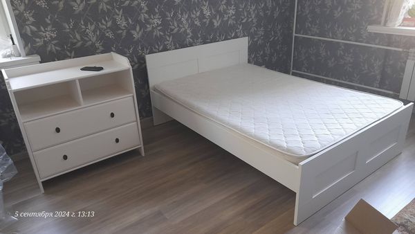 Собран новый белый комод с двумя выдвижными ящиками и двумя полками. Установлены ручки, проверена устойчивость, размещен рядом с кроватью на ламинированном полу в спальне.