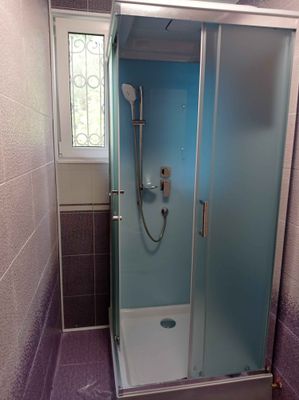 Изображена современная душевая кабина в ванной комнате с фиолетовой плиткой. Установлены стеклянные двери, душевой шланг и смеситель. Окно с решеткой обеспечивает естественное освещение.