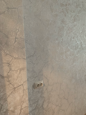 Стены оклеены обоями с текстурным узором, создающим эффект мрамора. Поверхность ровная, стыки аккуратные, розетка интегрирована без зазоров. Работа завершена качественно.